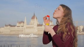 Beautiful girl blows bubbles. Attractive young woman blowing bubble in the Budapest. Close up shot of blonde female on the city view. Sunny spring day, woman having fun.