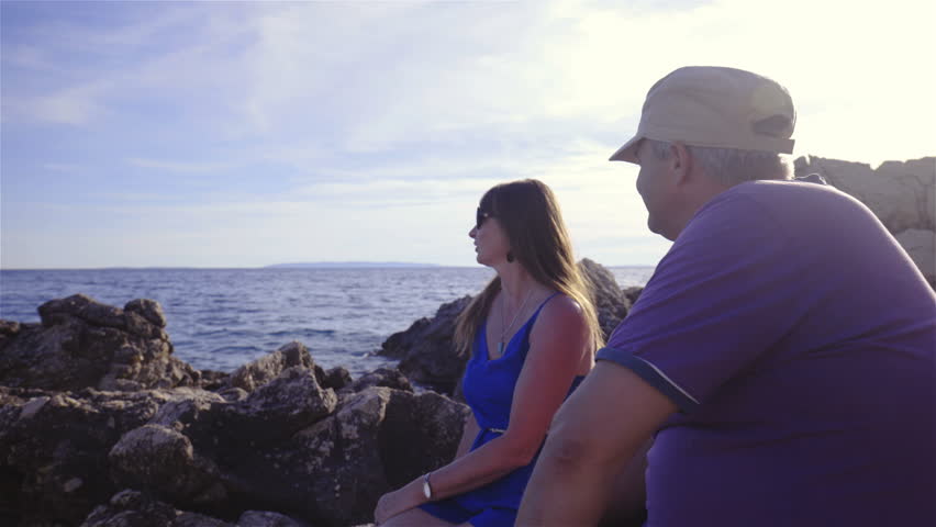 Middle age couple siting on rocks at sea 4K. Dolly slide shot of two person in focus with sunshine in background and sea behind rocks.