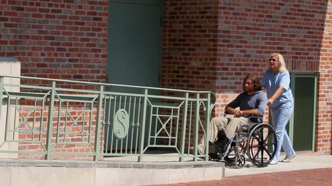 Female nurse in uniform pushes a man patient up a handicapped ramp to a building entrance.