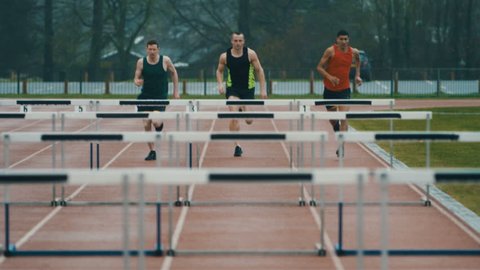 Competitive athletes running & jumping over hurdles at athletics track in slow motion