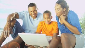 Young happy ethnic parents and son using laptop for online chat on the beach 