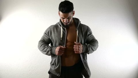 Bodybuilder handsome man undressing, taking off jacket against white background, revealing muscular naked torso in studio shot.