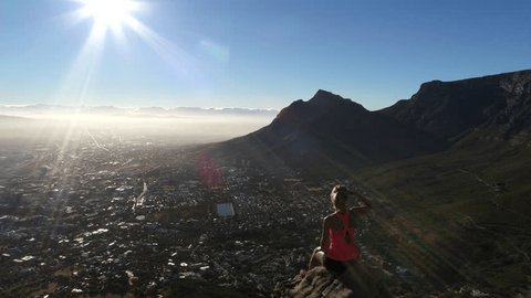 Stunning aerial shot of a young woman sitting high up on a rock looking down at Cape Town downtown surrounded by mountain landscape.
Drone shot of a woman hiker sitting on mountain top 
