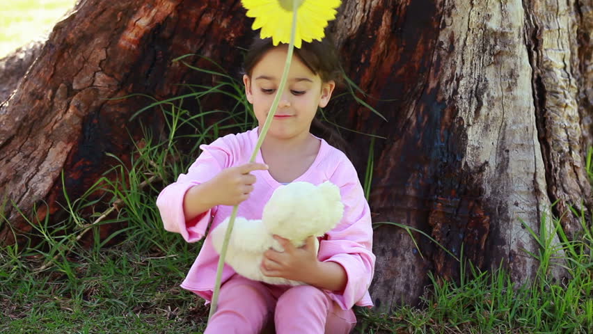 Smiling girl playing with a flower and a teddy bear in front of a tree in a park