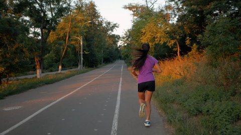 Girl runs through the park in the morning. Sporty young attractive brunette woman running on empty road in the morning. Slow motion. Back view.