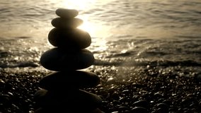 Closeup of pyramid made by sea stones on stony seashore at blurred golden light sea water background. Beautiful nature landscape with splashing waves of dark water. Real time video footage