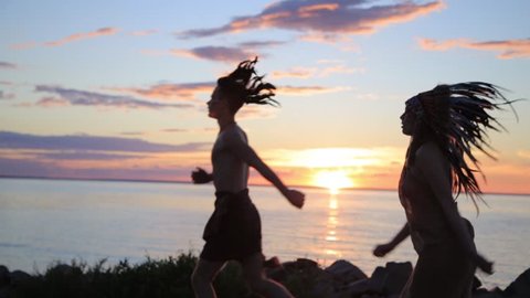 Silhouettes of running man and woman running jogging on the sea beach at sunset. Native american outfit and accessories.