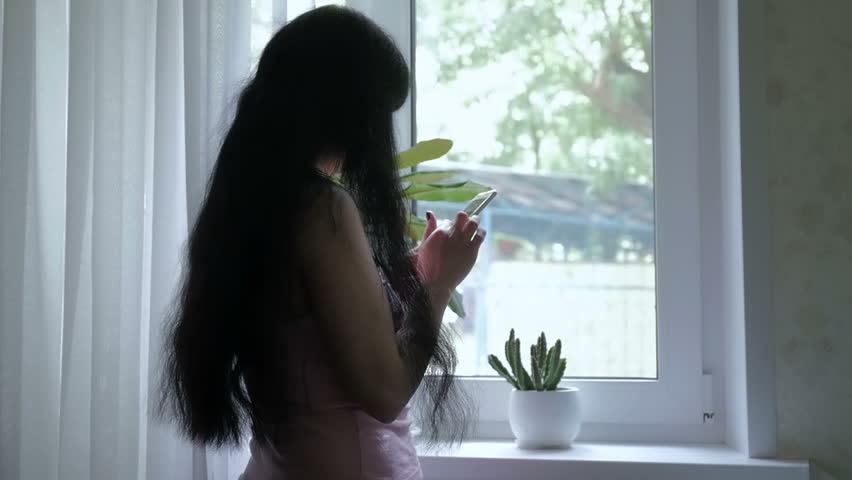A young, attractive girl with long beautiful hair is standing by the window and talking on the phone, writes sms. Slow motion. Sun | Shutterstock HD Video #29062840