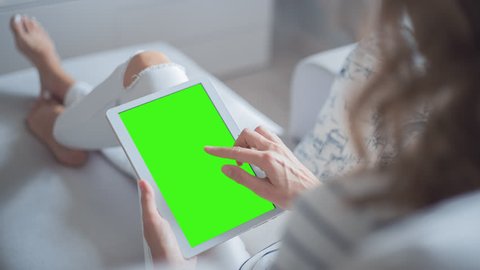 Young Woman in white jeans sitting on couch uses Tablet PC with pre-keyed green screen. Few types of gestures - scrolling up and down, tapping, zoom in and out. 