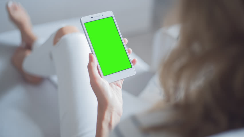 Young Woman in white jeans sitting on couch uses SmartPhone with pre-keyed green screen. Few types of gestures - scrolling up and down, tapping, zoom in and out.  Royalty-Free Stock Footage #29065318