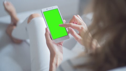 Young Woman in white jeans sitting on couch uses SmartPhone with pre-keyed green screen. Few types of gestures - scrolling up and down, tapping, zoom in and out. 
