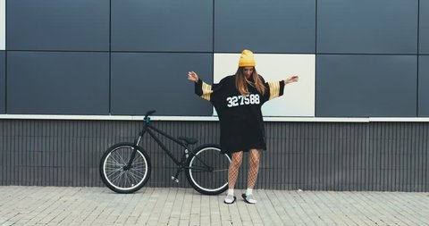 Young stylish Caucasian female dancing near a bicycle against grey wall background. 4K UHD RAW edited footage
