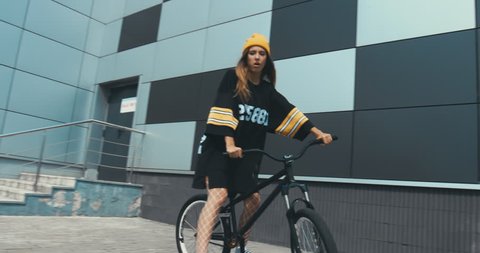 Young stylish Caucasian female in hockey style jersey riding her bicycle against grey wall background. 4K UHD RAW edited footage