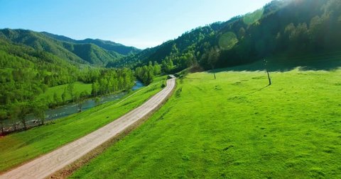 Aerial UHD 4K view. Low air flight over mountain rural dirt road and meadow at sunny summer morning. Near green trees, sun rays and mountain river