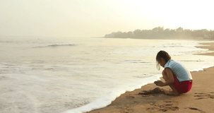 Video footage of a little girl with casual clothes, playing sand alone on the tropical beach in the morning. Shot in 4k resolution
