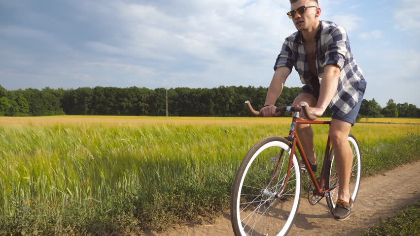 Young man riding vintage bicycle at the rural road over field. Sporty guy cycling along country trail outdoor. Male cyclist riding bike in the countryside. Healthy active lifestyle