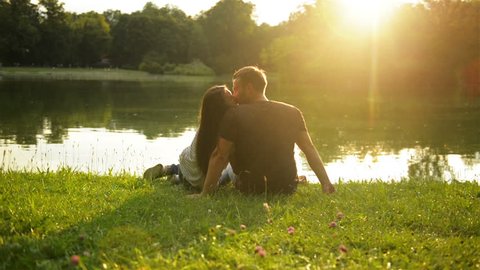 Two Lovers Kissing During Beautiful Sunset Sitting near the Lake in the City Park. Back View of Happy Couple Outside.