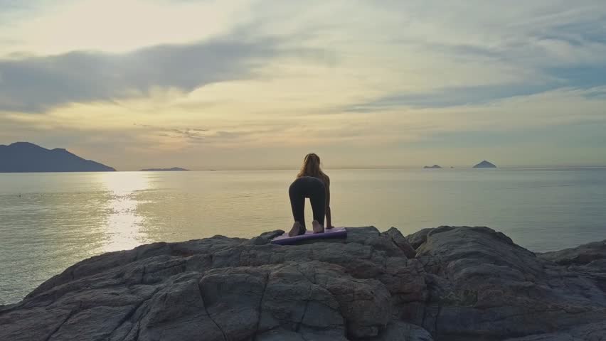 backside view young girl stands on knees in yoga pose on rocky beach against boundless ocean and pictorial sunrise