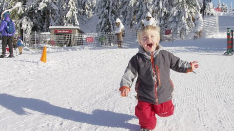 A young boy runs towards the camera with people and snow covered trees behind him.  Camera starts close to him. Near Santa's Workshop on the top of Grouse Mountain. 4k. Slow Motion.
