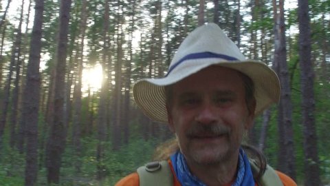 Tired but happy tourist in a hat with a backpack in a pine forest on a summer evening