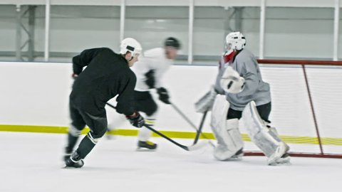 Tracking shot of ice hockey forward in black gear dribbling puck, then passing it to teammates in white uniform who shooting it and scoring goal