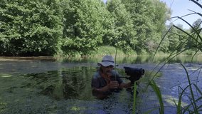 Mature male videographer in water with homemade camera shoot birds and insects on river among green cane