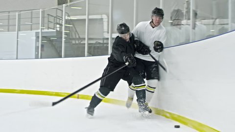 Slow motion tracking of hockey forward in white uniform dribbling puck along ice rink board, then falling as player from opposing team boarding him
