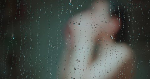Woman Showering Focus on Water Drops on Door Move Right. a woman from chest up is blurred showering and turning while shot moves right focus on water drops on glass door falling in slow motion
