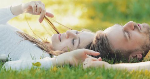 Happy Couple Relaxing on a Green Grass, Dolly Shot. SLOW MOTION 4K. Smiling man and woman in love are talking, enjoying summertime. Joyful family. Young lovers lying on a lawn, relaxing outdoors.