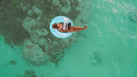 Aerial drone quad copter footage of woman using laptop computer, concept of tourist woman floating on lilo inflatable and working on laptop computer in tropical exotic ocean. Remote working anywhere.