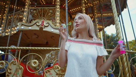 Beautiful young blonde girl blowing soap bubbles. Gorgeous young teen girl blowing bubbles. Outdoor summer portrait of attractive happy woman having fun in amusement park. Slow motion.