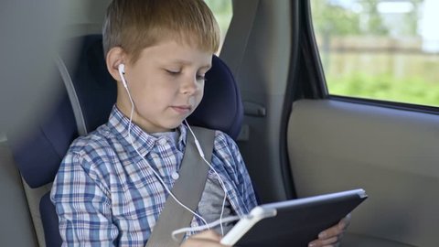 Little boy sitting in car seat and watching something on digital tablet with headphones while traveling in car
