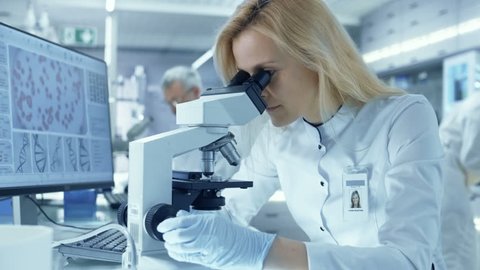 Female Research Scientist Looks at Biological Samples Under Microscope. She and Her Colleagues Work in a Big Modern Laboratory/ Medical Center. Shot on RED EPIC-W 8K Helium Cinema Camera.
