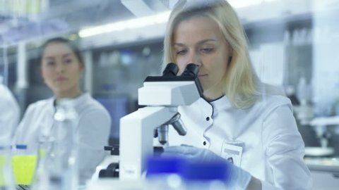 Female Research Scientists Passes Slided to Her Colleague Who Observes Sample under Microscope. They Work in a Big Research Center/ Laboratory. Shot on RED EPIC-W 8K Helium Cinema Camera.