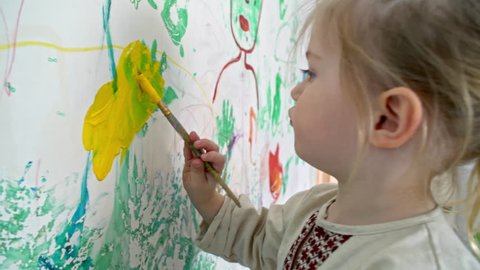Handheld side view of little boy with ponytail holding small paintbrush and drawing on wall of his room with yellow gouache