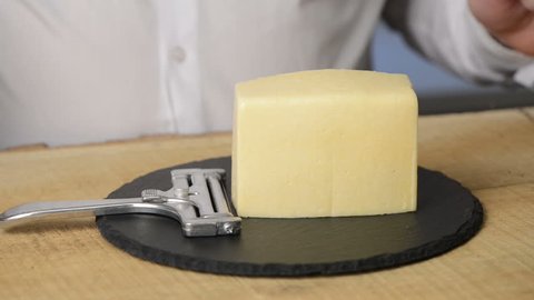 The man cuts cheese into thin slices with use of a cheese slicer on a black round chopping board from slate