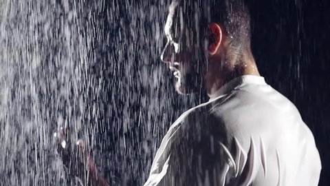 portrait of brutal man with a beard and broad shoulders in a white shirt. he stands in the rain and thought. stretched out his hand to catch the drops for meditation presence