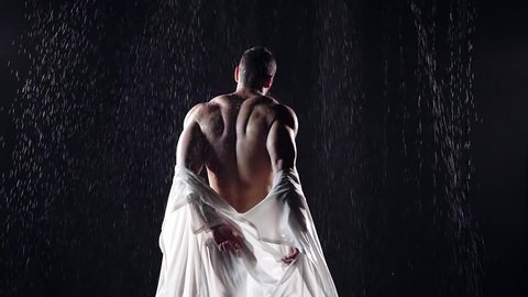 sports man's muscular back standing in the rain at night. he takes off his wet shirt in white. muscles play on the backlight