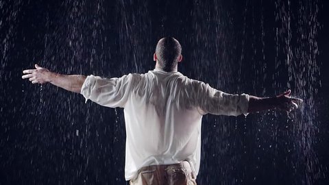 view from the back. average plan. a large man in white clothes standing under a large rain threw his arms to the sky and pray to God