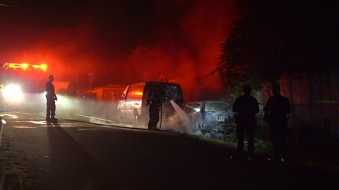 Firefighters douse a vehicle that was fully engulfed in flames as police officers stand near by and assess the scene at night.
