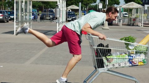 A young man is riding a trolley in a parking lot near a supermarket. The basket is full of products. Fun and good mood. Merry shopping. Dad buys food