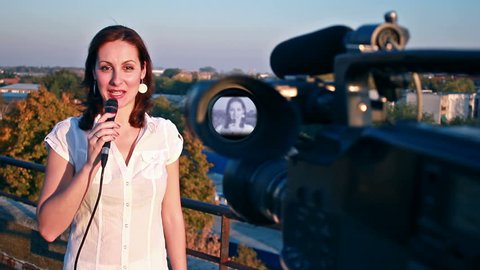 TV reporter on assignment ; TV reporter in front of the camera gives notice of the weather forecast at the top of the building