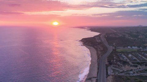 Aerial timelapse in motion (hyperlapse) shot from above at sunset with a drone over the Pacific ocean, Pacific Coast Highway (PCH) with traffic and beach with pink, orange and red skies at twilight.