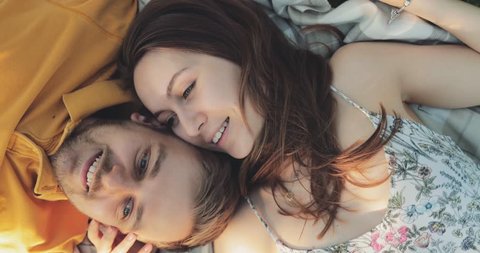 Happy Couple Relaxing on a Green Grass, Top View. SLOW MOTION 4K. Smiling man and woman in love are talking, enjoying summertime. Joyful family. Young lovers lying on a lawn, relaxing outdoors.