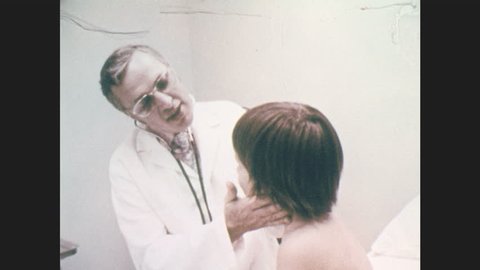 1970s: UNITED STATES: male doctor listens to boy's chest in clinic. Girl and mother talk with doctor in clinic