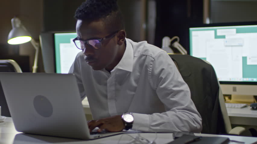 Handheld camera shot of concentrated black office worker working on laptop in the office at night