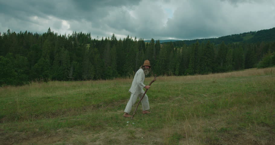 The full-length side view of the old man in old clothes, with long beard walking along the green mountains with the help of the cane.
