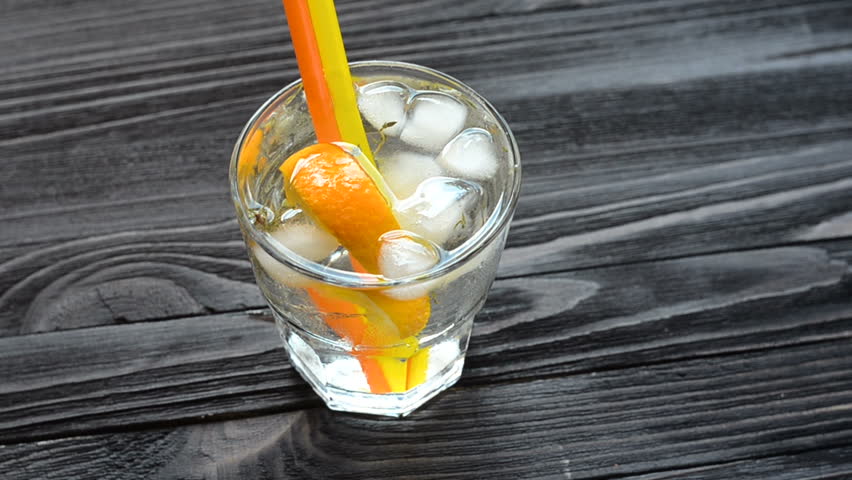 Hand stirs with straws a cocktail with an orange slice, ice and rosemary on dark wooden table