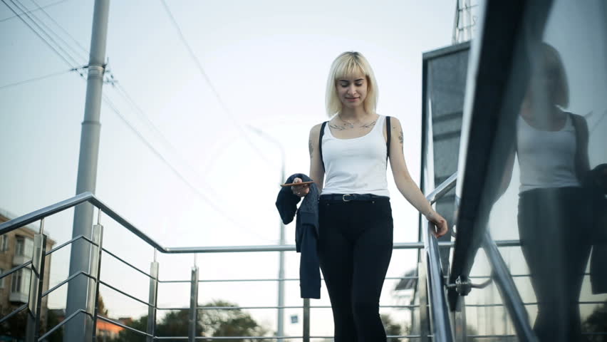 A young blond woman walks down the stairs in the city. Moves towards the camera.