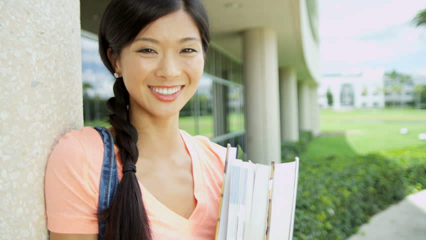 Portrait of happy young Asian American female student holding workbook standing before modern block college campus 